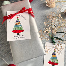 Xmas themed ribbon tag featuring a white background with a colourful xmas tree and a merry christmas wish on top with names at the bottom and tied to a gift with a red ribbon