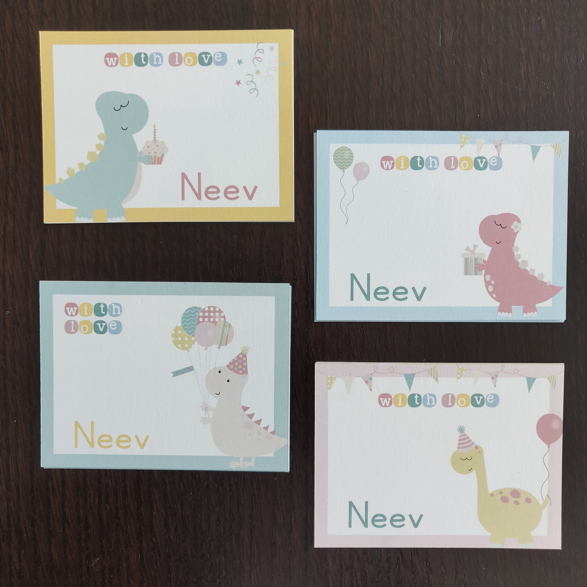Four personalized dinosaur-themed greeting cards arranged in a 2x2 grid on a dark wood surface. Each card has a different pastel border color (yellow, light blue, light teal, and light pink) and features a cute cartoon dinosaur with balloons, a party hat, and the name 'Neev' printed below.